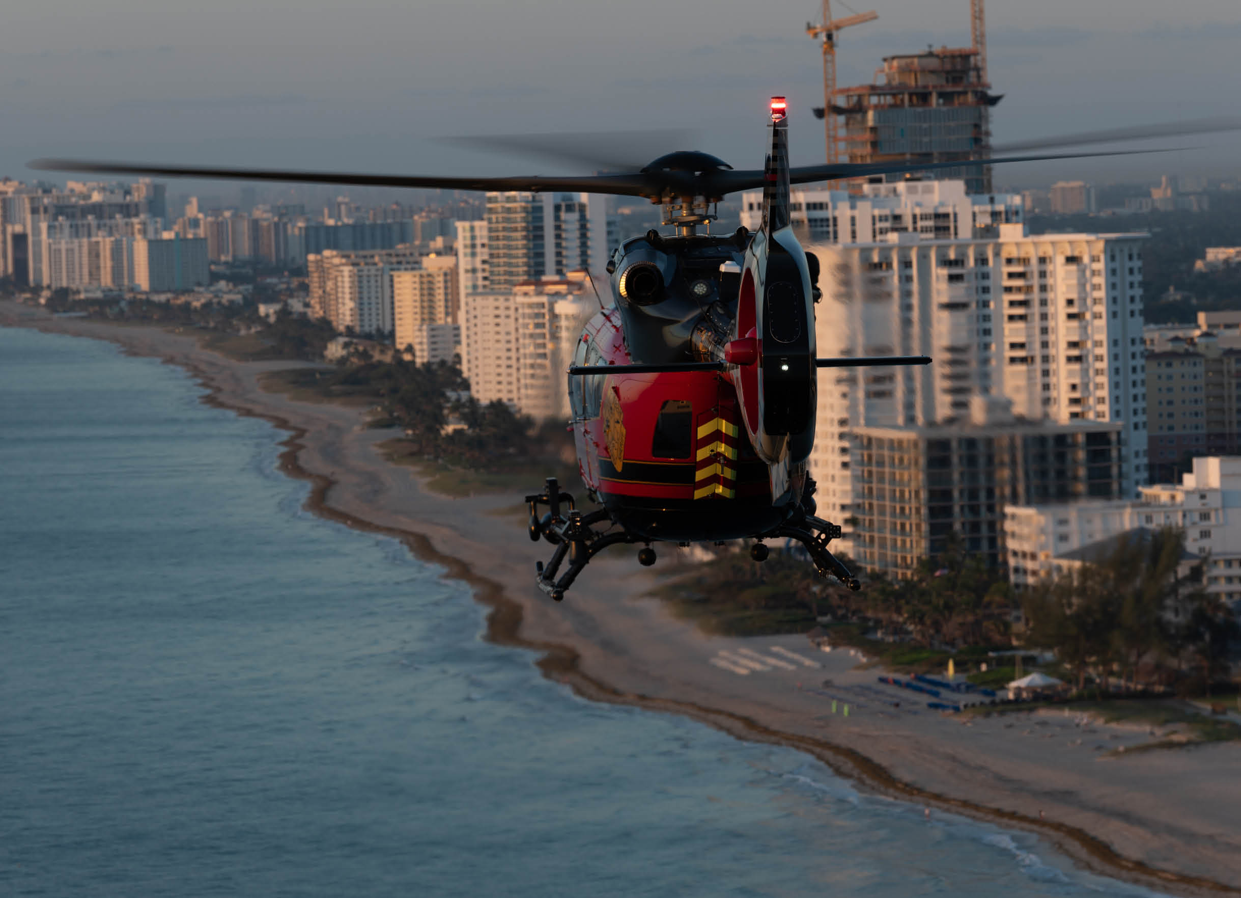 A red and black helicopter is flying over a beach near a city. AI generated content