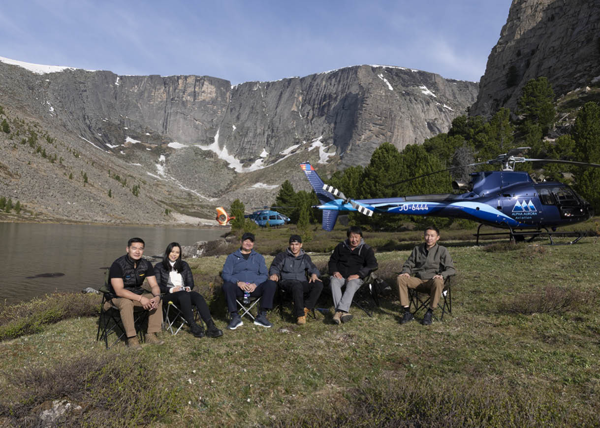 A group of people are sitting in chairs on a grassy field, posing for a picture. There is a helicopter in the background, and a lake nearby. AI generated content