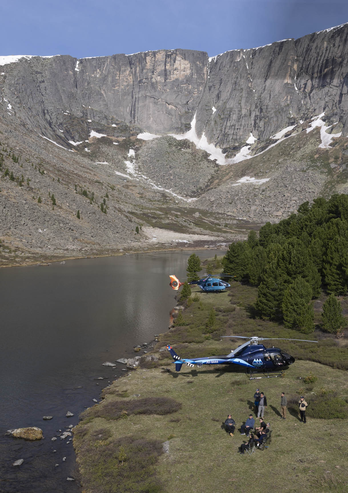 A group of people are standing on a grassy field near a lake. There are two kites in the air, one near the center of the field and the other closer to the right side. A blue car is parked on the grass, and a backpack is placed nearby. The scene appears to be a recreational gathering with people enjoying the outdoors. AI generated content