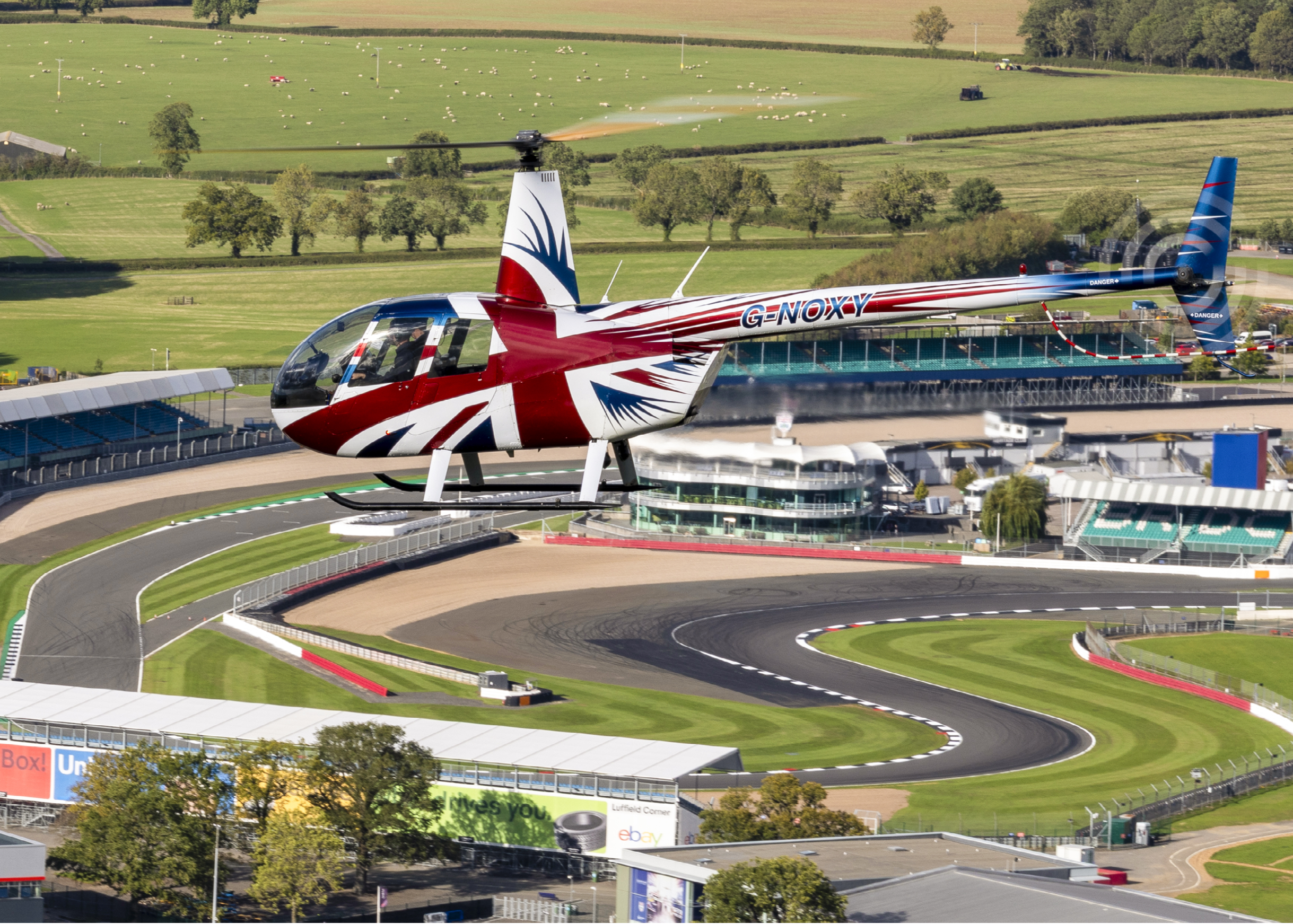 A British Airways jet is flying low over a race track with a red, white, and blue design. The airplane is flying over a group of cars on the track. There are also several people on the track, likely spectators or participants in the race. The scene is set in a rural area with a grassy field and a building nearby. AI generated content