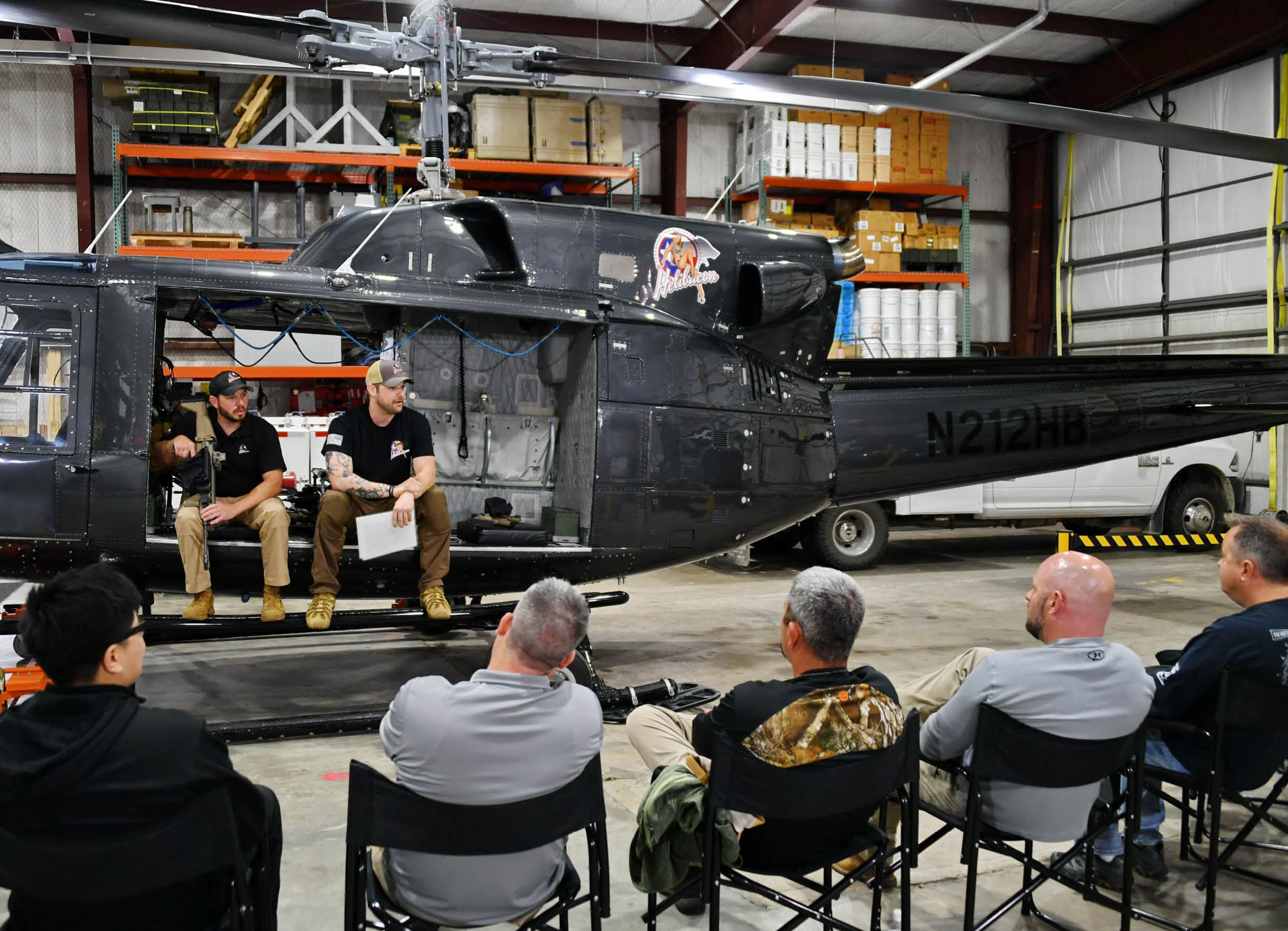 A group of people are gathered around a helicopter, with some sitting on chairs and others standing. The helicopter is parked inside a building, and there are several chairs and a truck visible in the scene. AI generated content