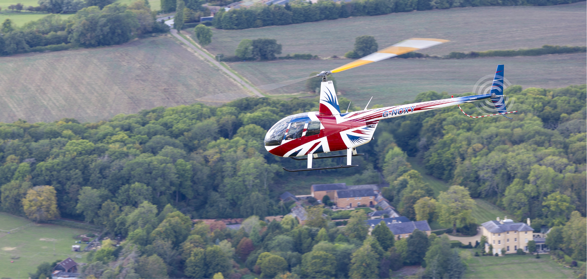 A red and white helicopter with a British flag on it is flying over a field. AI generated content