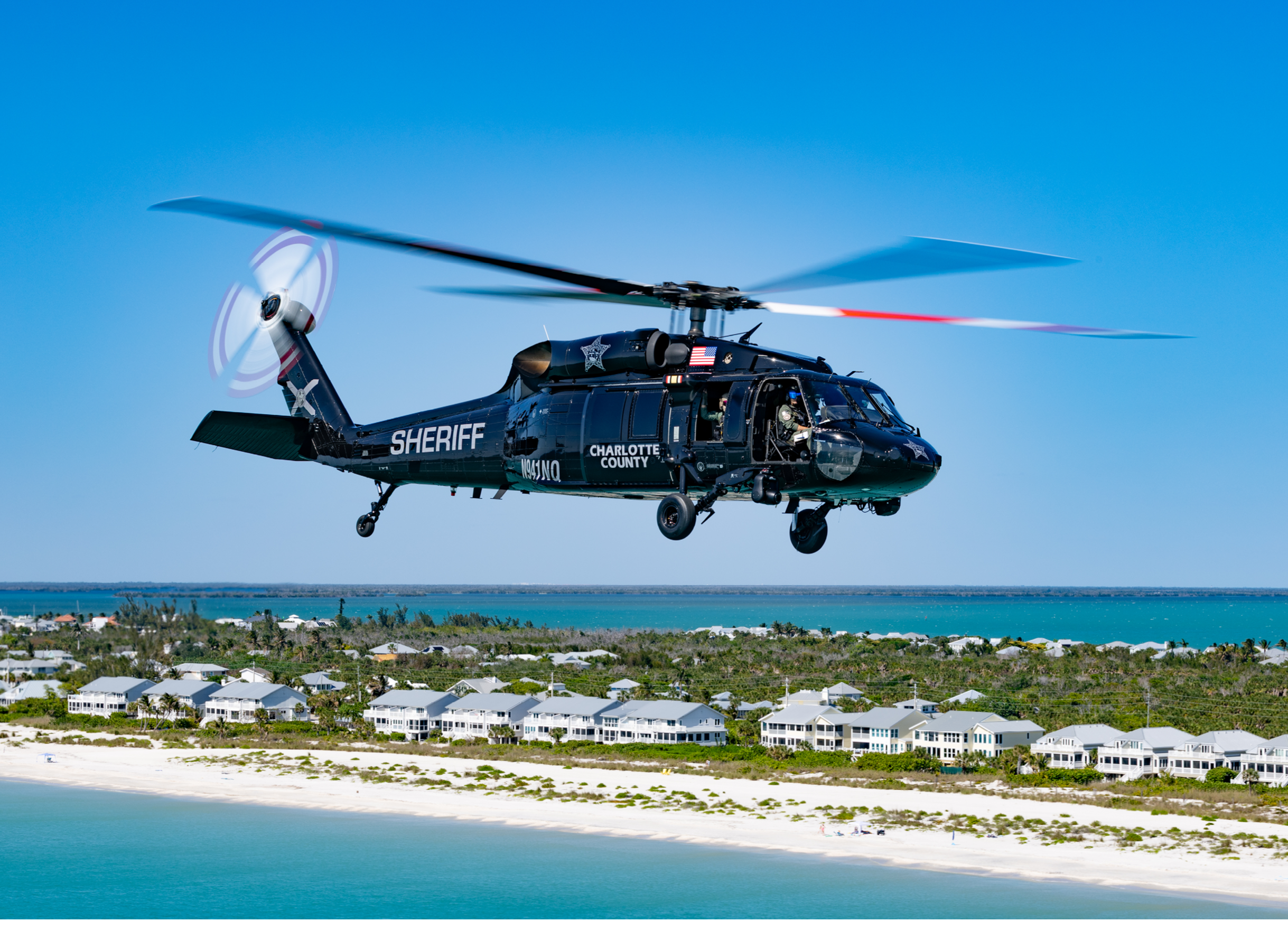 A black helicopter flying over a beach. Description generated by AI