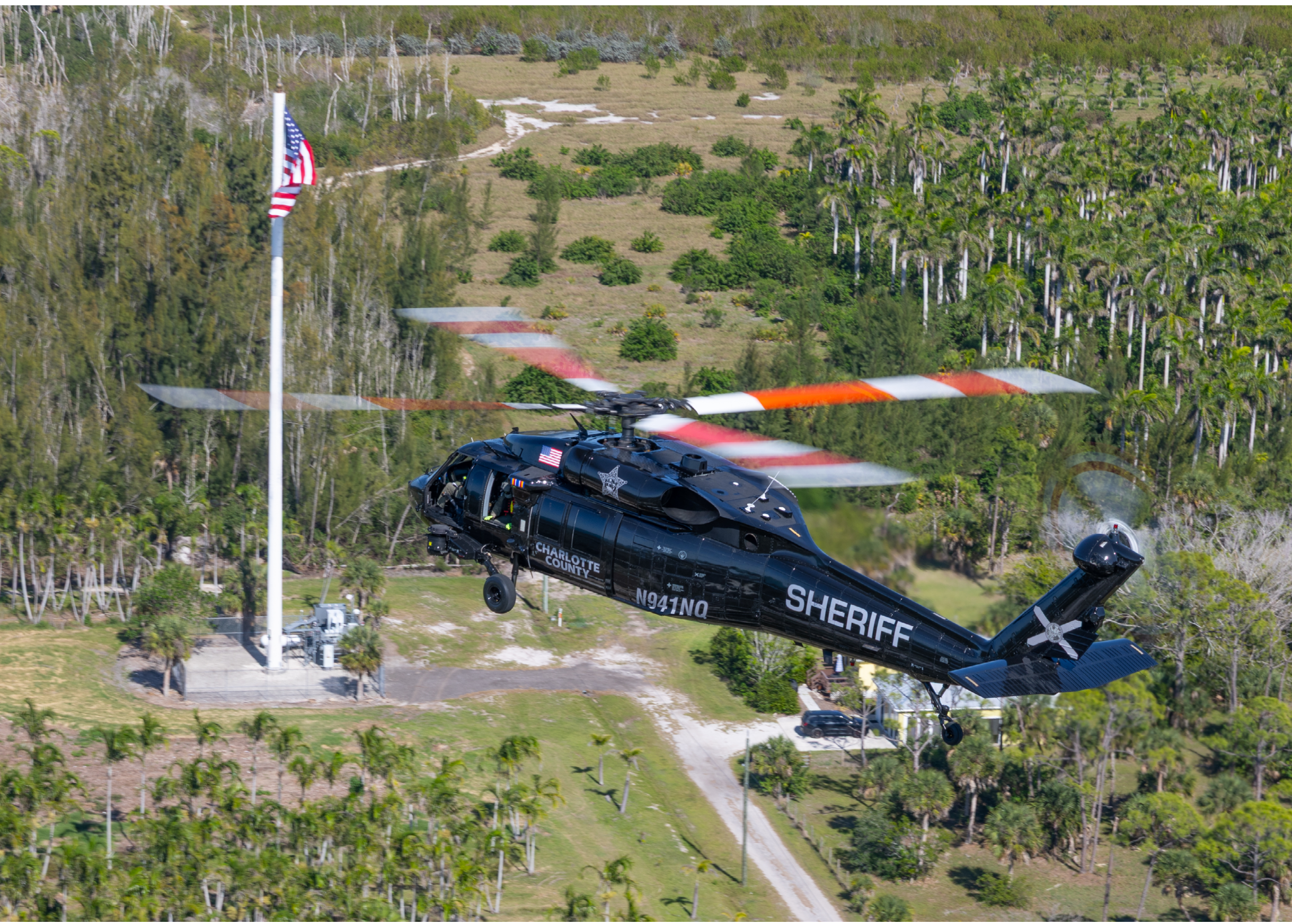 A black helicopter with a red cross on it is flying over a field. Description generated by AI