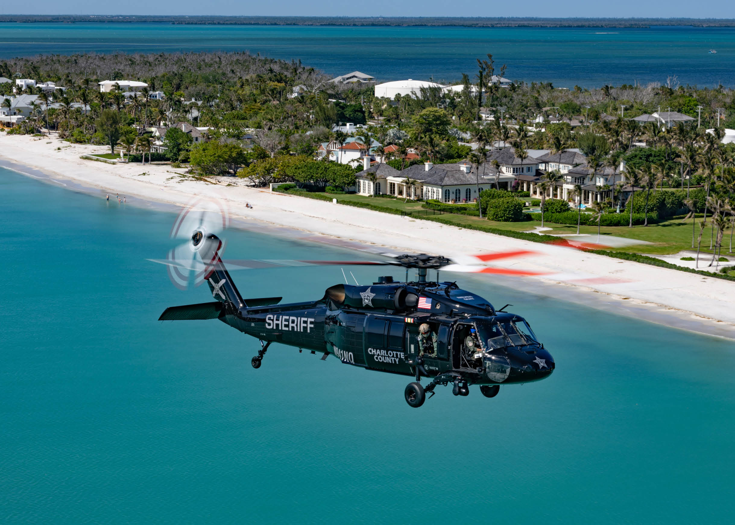 A black helicopter flying over a beach. Description generated by AI