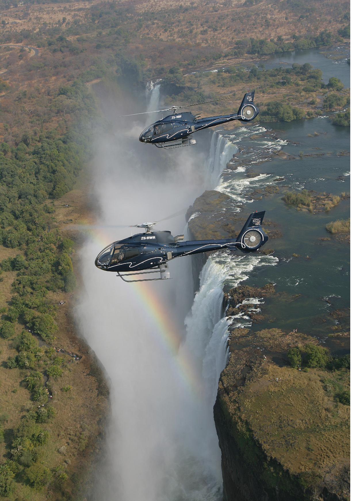Two helicopters flying over a waterfall. Description generated by AI