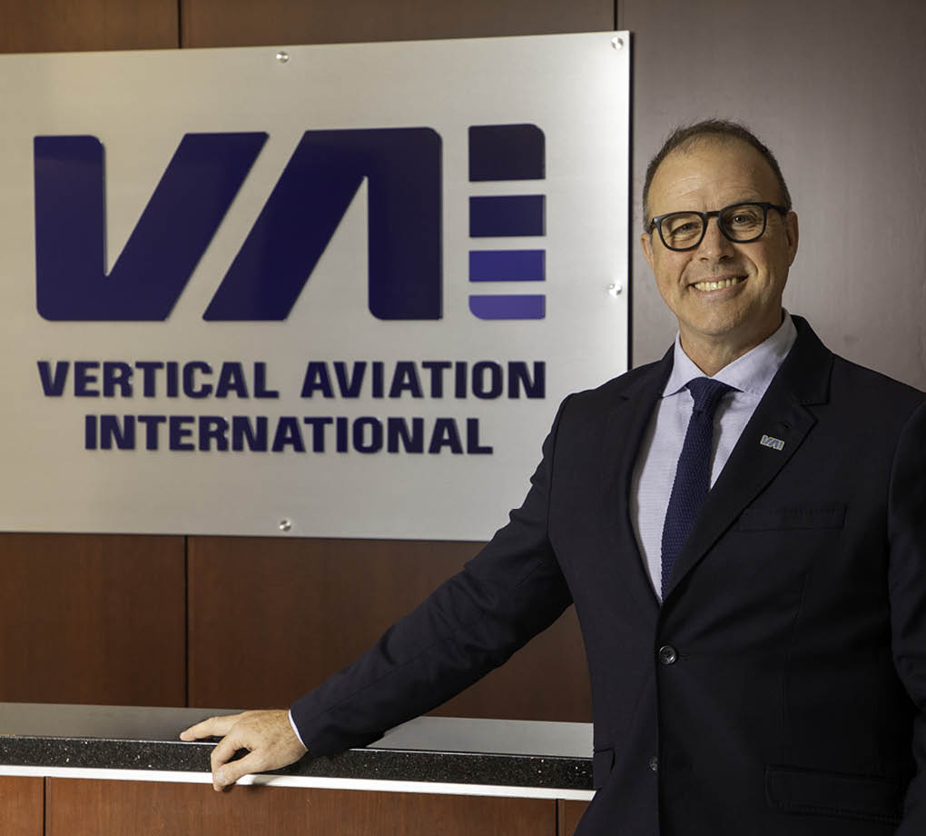 A man in a suit stands in front of a sign that says Vertical Aviation International. Description generated by AI