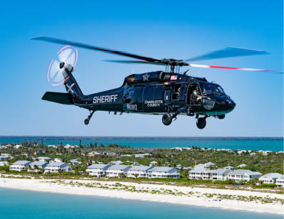 A black helicopter flying over a beach. Description generated by AI