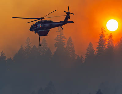 A helicopter flying over a forest during a sunset. Description generated by AI