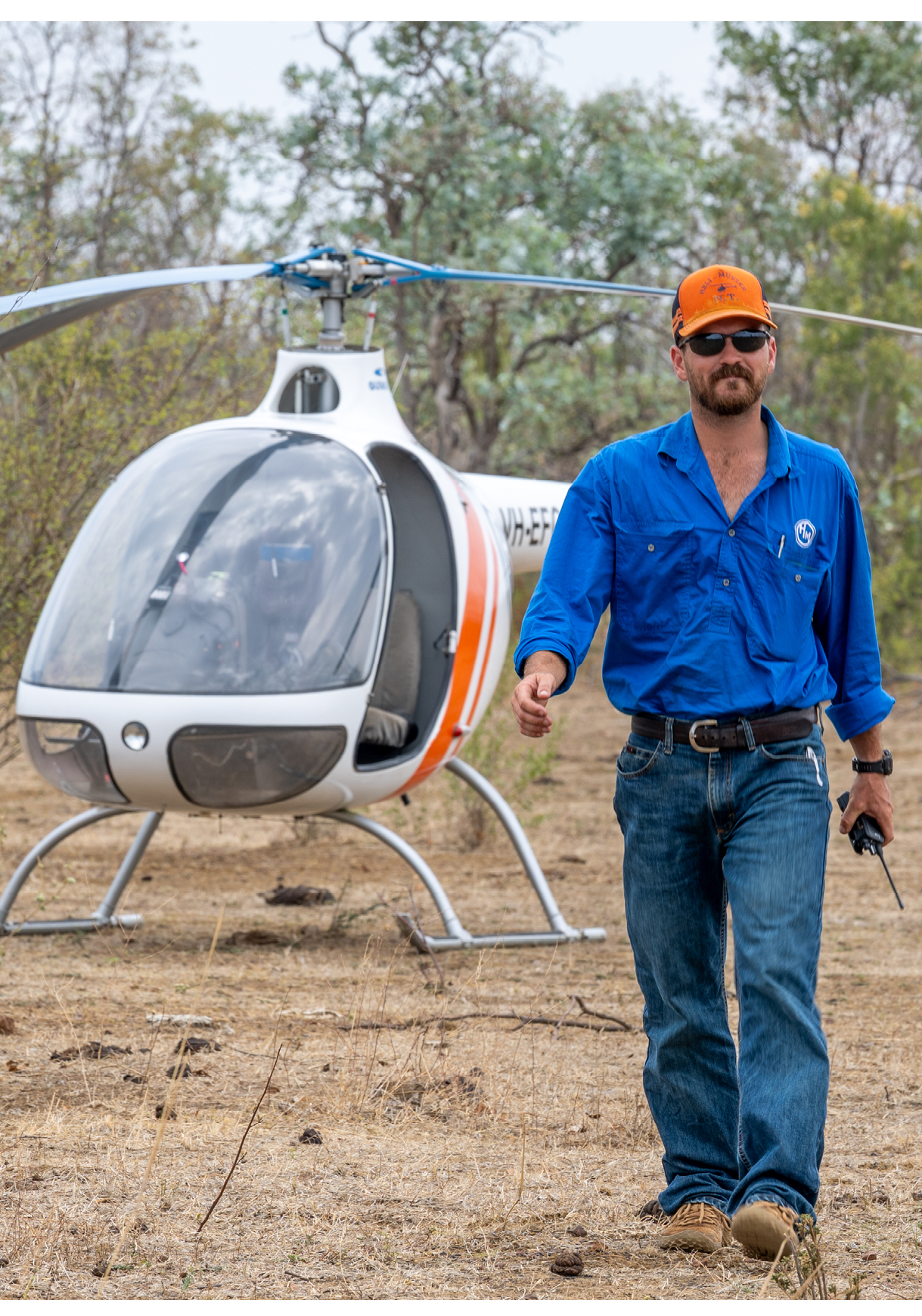 A helicopter is parked on dry, grassy ground near sparse trees with thin branches and small leaves. Description generated by AI