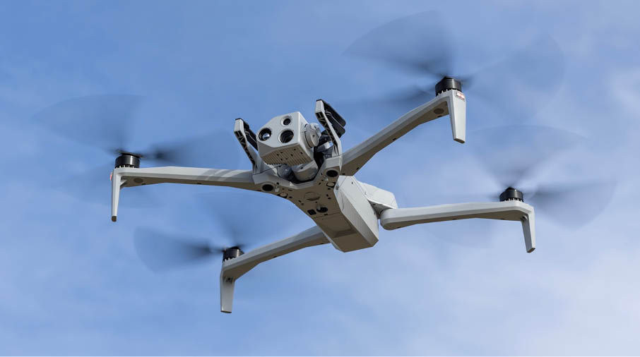 A white drone with four spinning propellers is flying against a clear blue sky. Description generated by AI