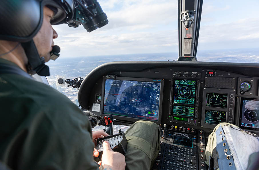 A person wearing a helmet and headset is operating the controls inside a helicopter cockpit with multiple digital displays showing flight information. Description generated by AI