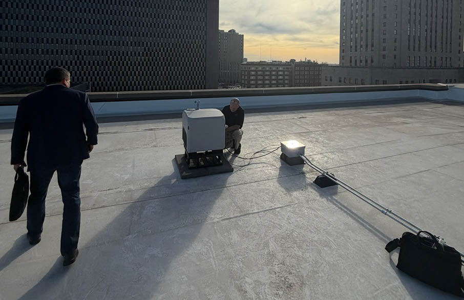 Two people stand on a rooftop with large buildings in the background under a partly cloudy sky with the sun low on the horizon. Description generated by AI