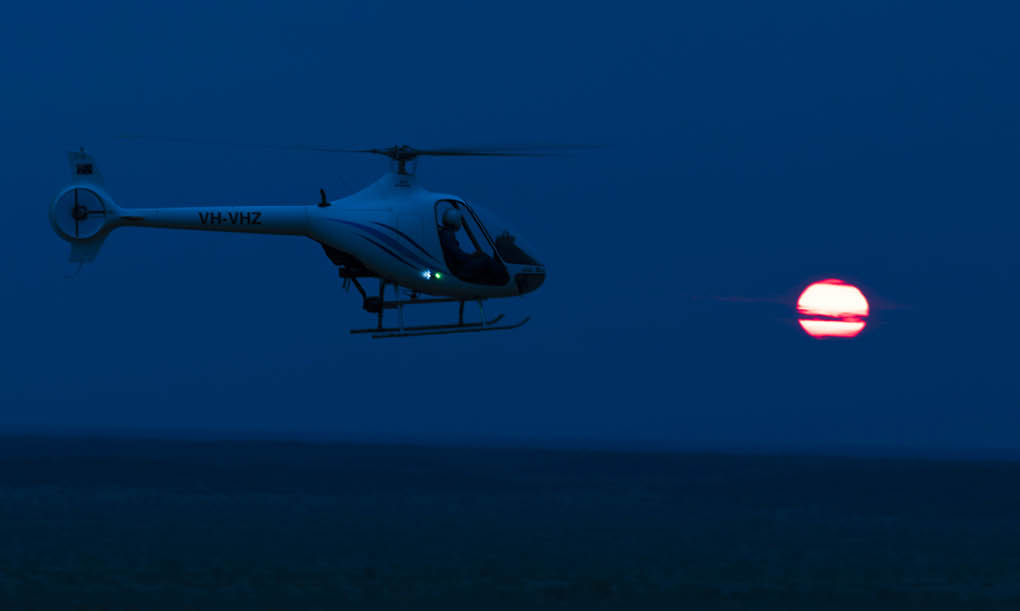 A helicopter is flying at dusk with a large red sun low on the horizon against a dark blue sky. Description generated by AI