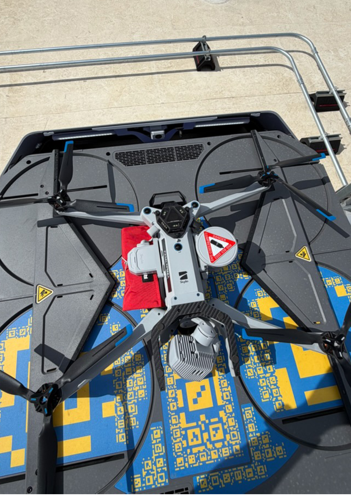 A large drone with four propellers is resting on a surface decorated with a blue and yellow pattern, with metal rods and a small vent visible in the background. Description generated by AI