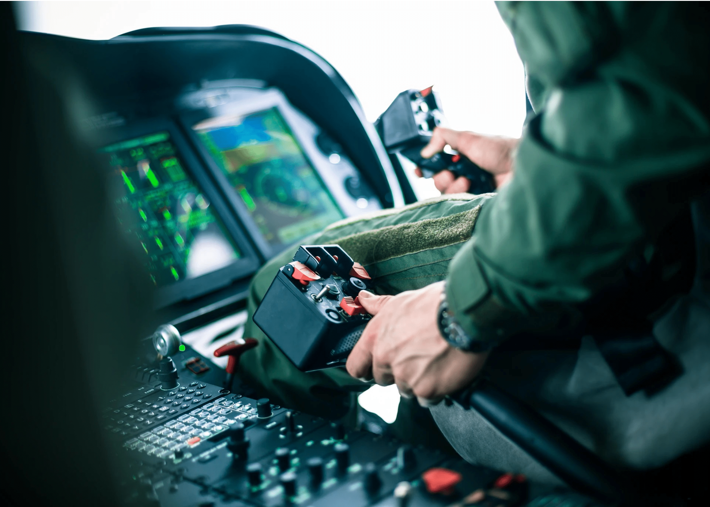 A person in a green flight suit is operating dual control sticks inside a cockpit with illuminated digital instrument panels. Description generated by AI
