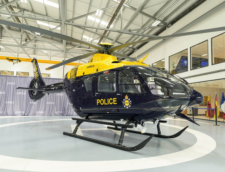 A dark blue and yellow police helicopter is parked inside a spacious, well-lit hangar with large windows and a high ceiling. Description generated by AI