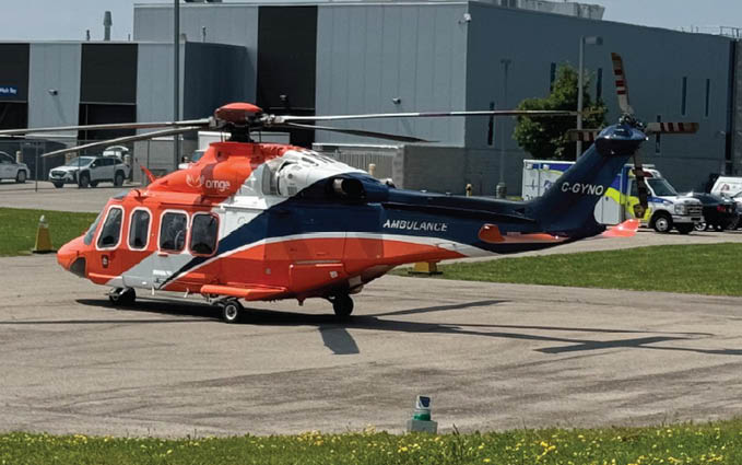 A helicopter with orange and blue markings is parked on a helipad surrounded by grass and a few traffic cones. Description generated by AI