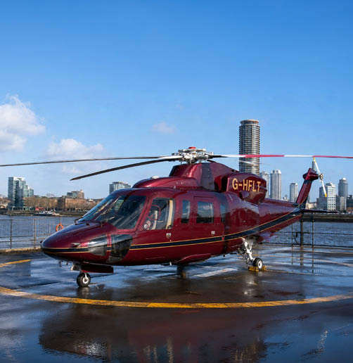 A red helicopter with the registration G-HFLT is parked on a wet helipad near a body of water with city buildings in the background under a clear blue sky. Description generated by AI