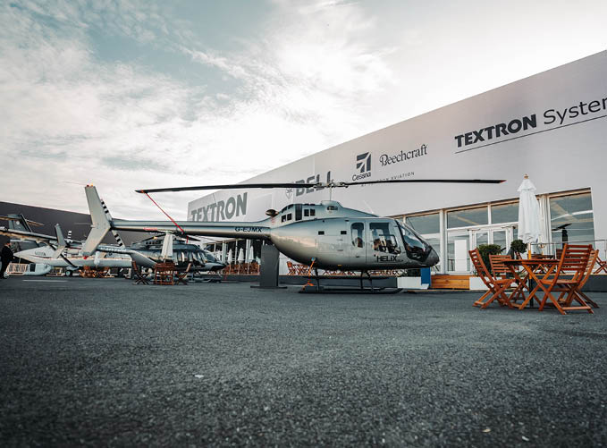 A helicopter is parked on a tarmac in front of a building with signs for Textron Systems, Beechcraft, and Bell, with outdoor wooden tables and chairs nearby. Description generated by AI