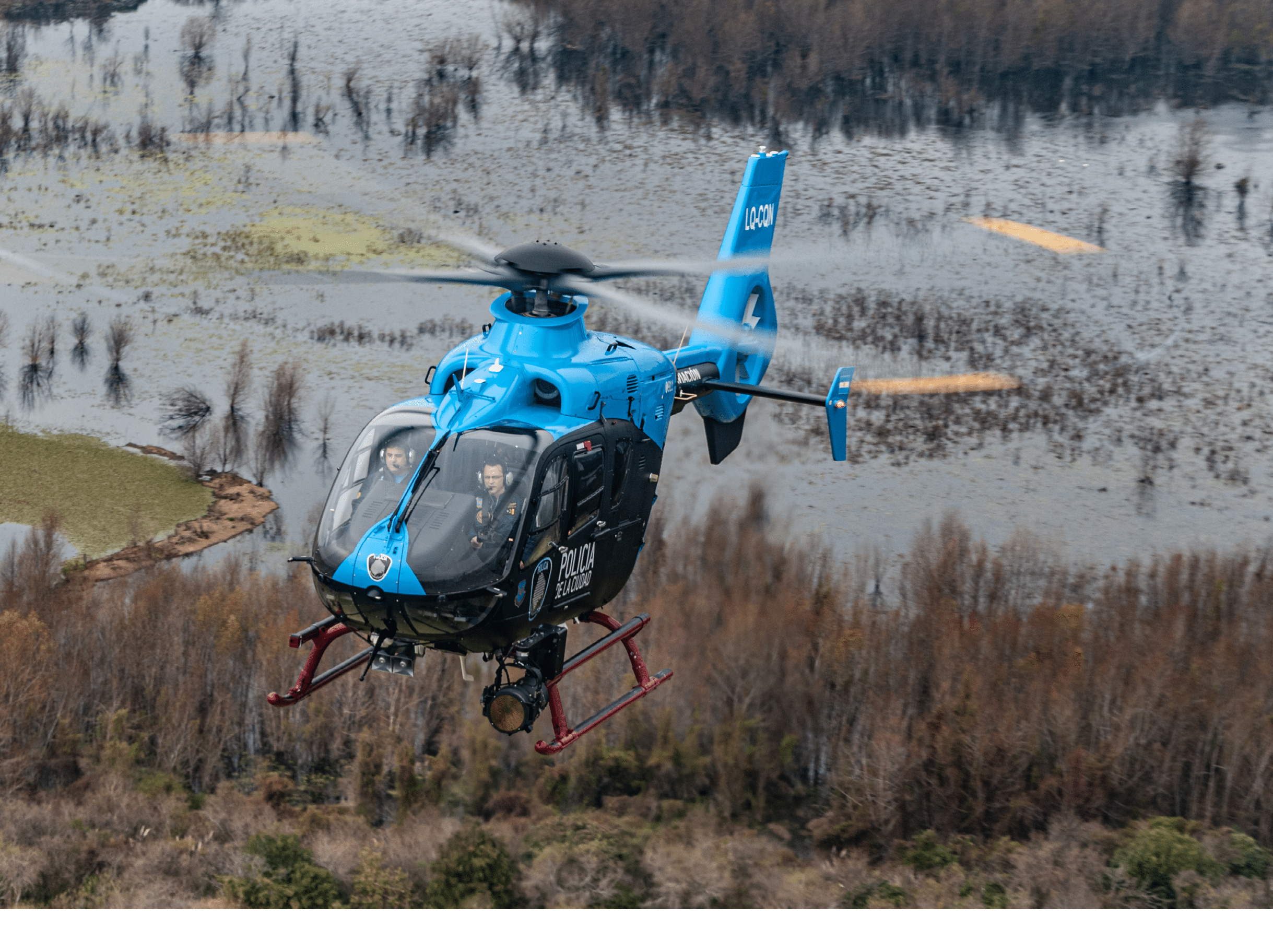 A blue helicopter is flying low over a marshy area with sparse trees and water. Description generated by AI