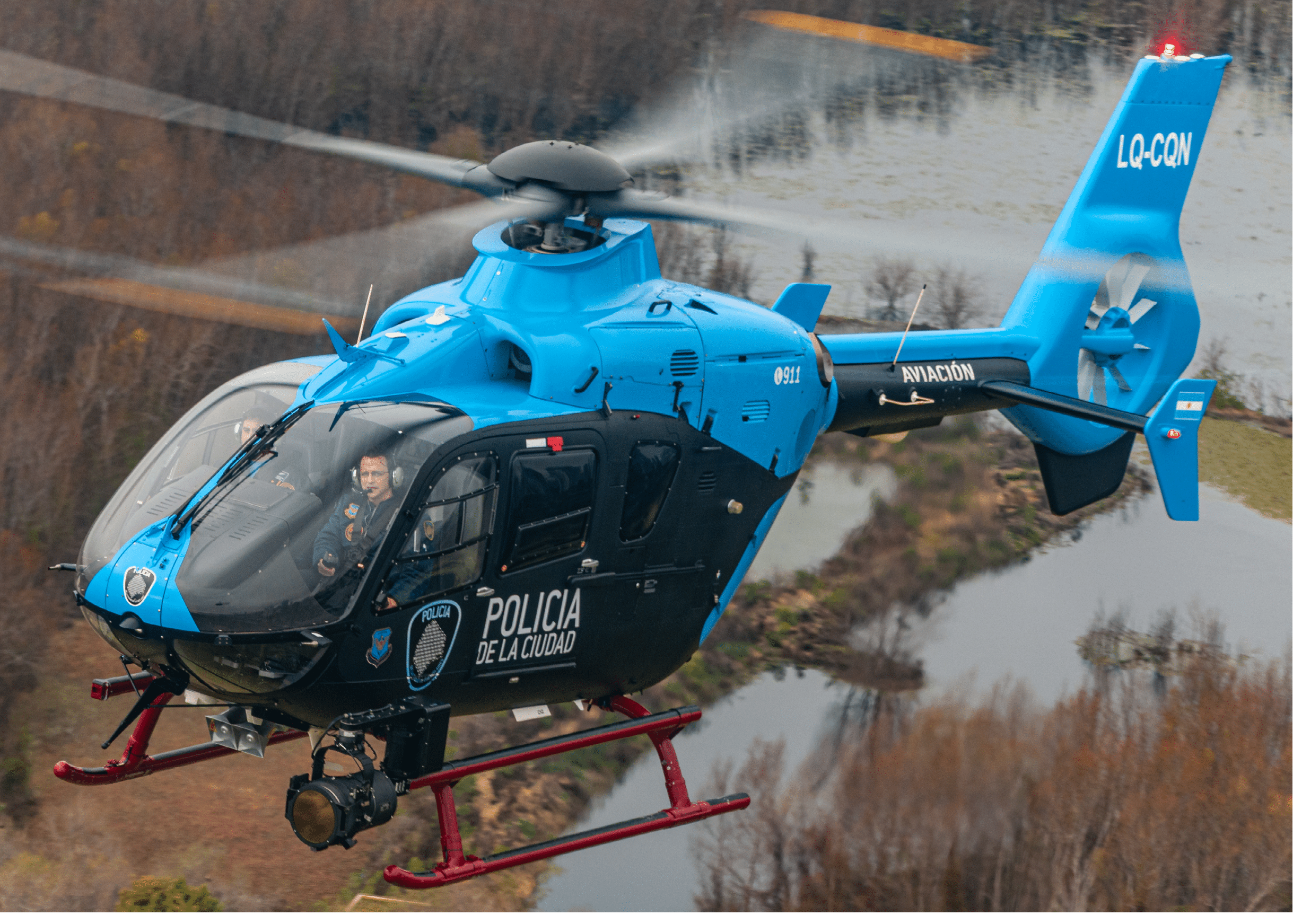 A blue and black police helicopter with the text “POLICIA DE LA CIUDAD" is flying over a river and wooded area. Description generated by AI