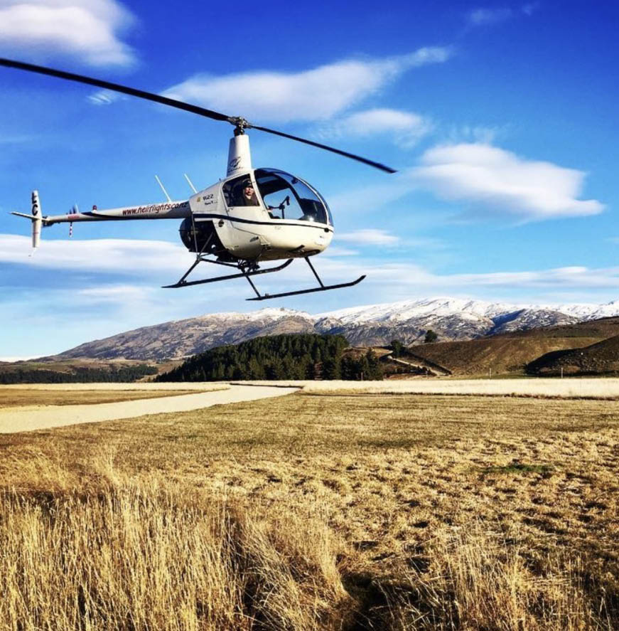 Two helicopters are flying low over a dry grassy field with mountains and a partly cloudy sky in the background. Description generated by AI