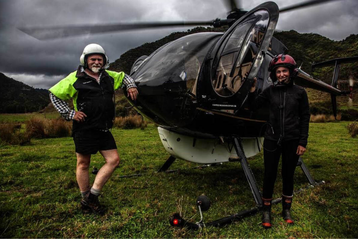 Two people wearing helmets and outdoor clothing stand on grass beside a small black helicopter under a cloudy sky. Description generated by AI