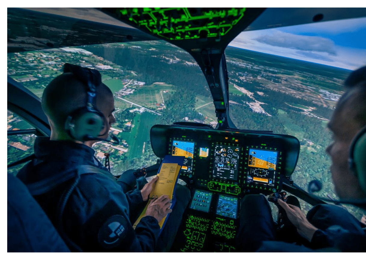 Two people wearing headsets are operating a helicopter cockpit with multiple digital displays showing flight information and an aerial view of the landscape below. Description generated by AI