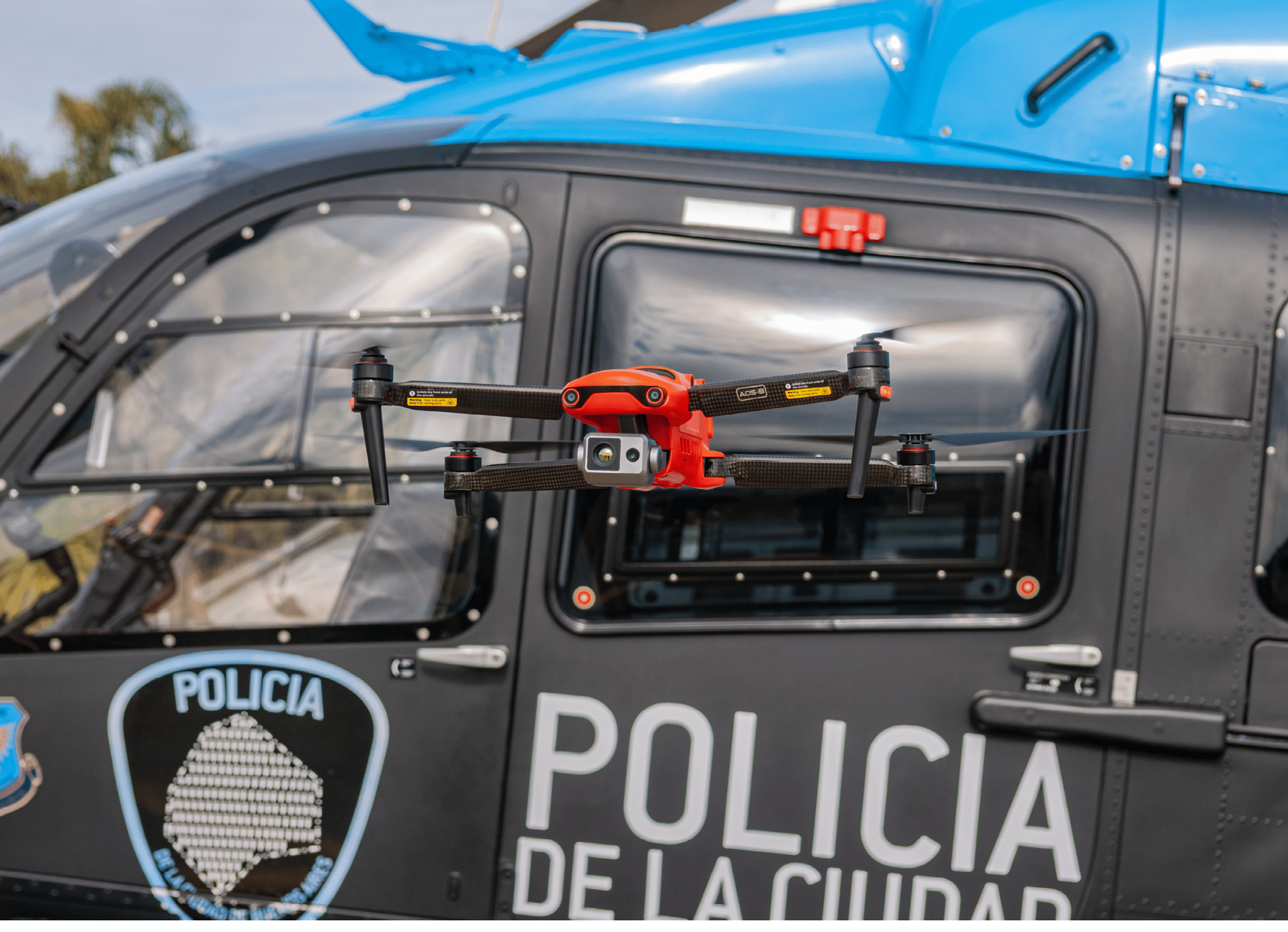 A red drone is flying in front of a black and blue police helicopter with the word “POLICIA" visible on the side. Description generated by AI