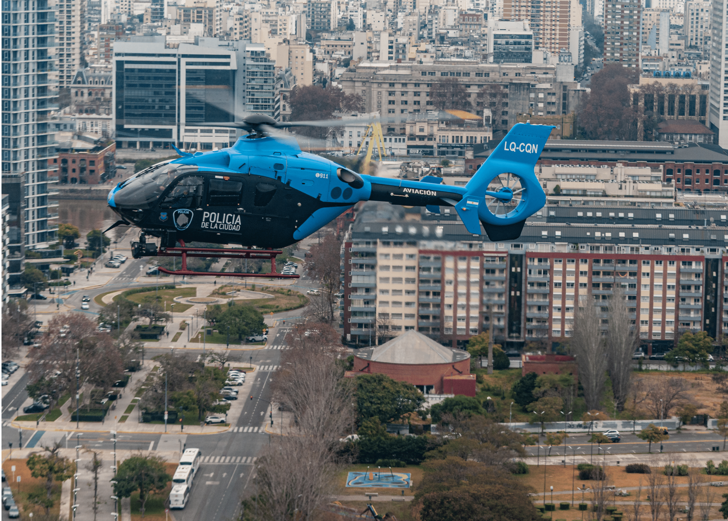 A blue and black police helicopter is flying over an urban area with multiple buildings and streets visible below. Description generated by AI