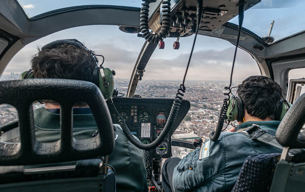 Two people wearing headsets are seated inside a helicopter cockpit with a cityscape visible through the windows. Description generated by AI