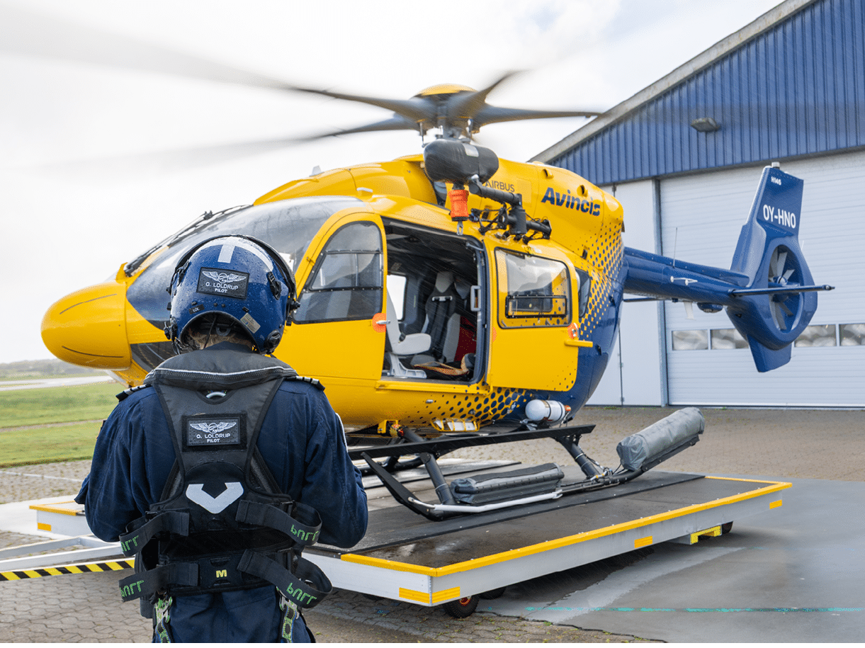 A person wearing a blue helmet and harness stands near a yellow helicopter with its door open on a platform outside a building. Description generated by AI