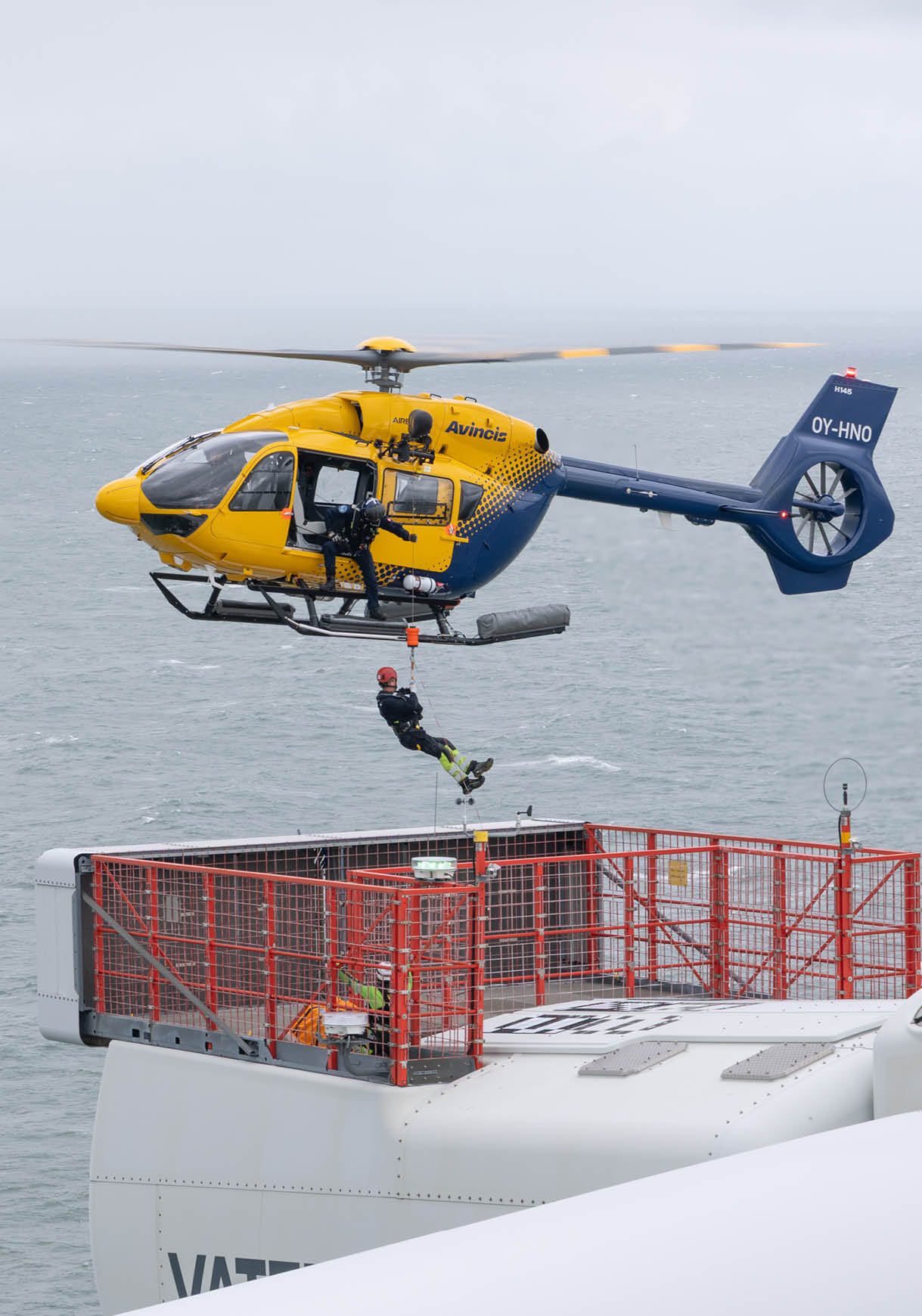A yellow helicopter is hovering near the edge of a ship above a gray, choppy sea under an overcast sky. Description generated by AI