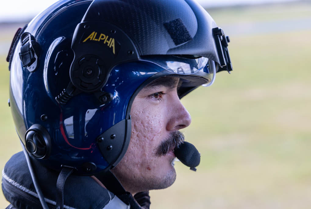 A person wearing a dark blue helmet with a microphone attached is looking to the right against a blurred outdoor background. Description generated by AI