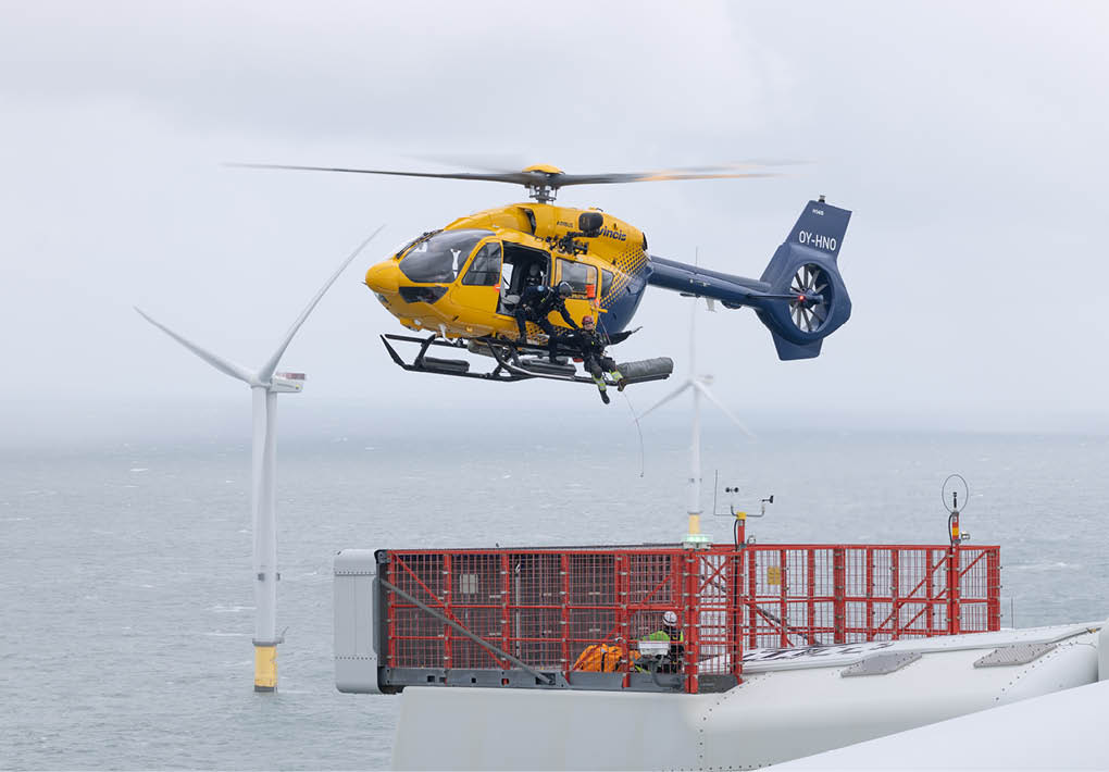 A yellow and blue helicopter hovers above a platform on a wind turbine with two people wearing safety gear visible on the platform and one person inside the helicopter. Description generated by AI