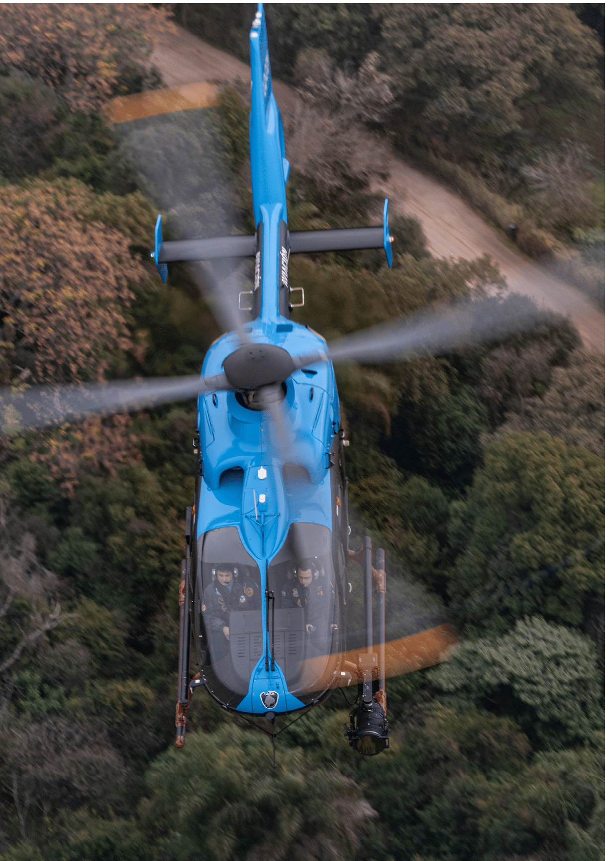 A blue helicopter is flying over a forested area with two people visible inside the cockpit. Description generated by AI