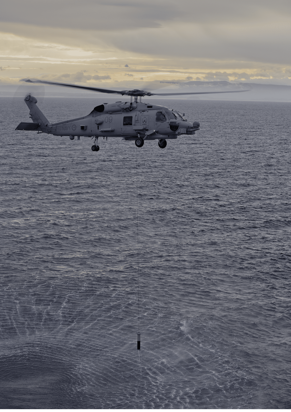 A military helicopter hovers above the ocean near a small floating object with a cloudy sky in the background. Description generated by AI