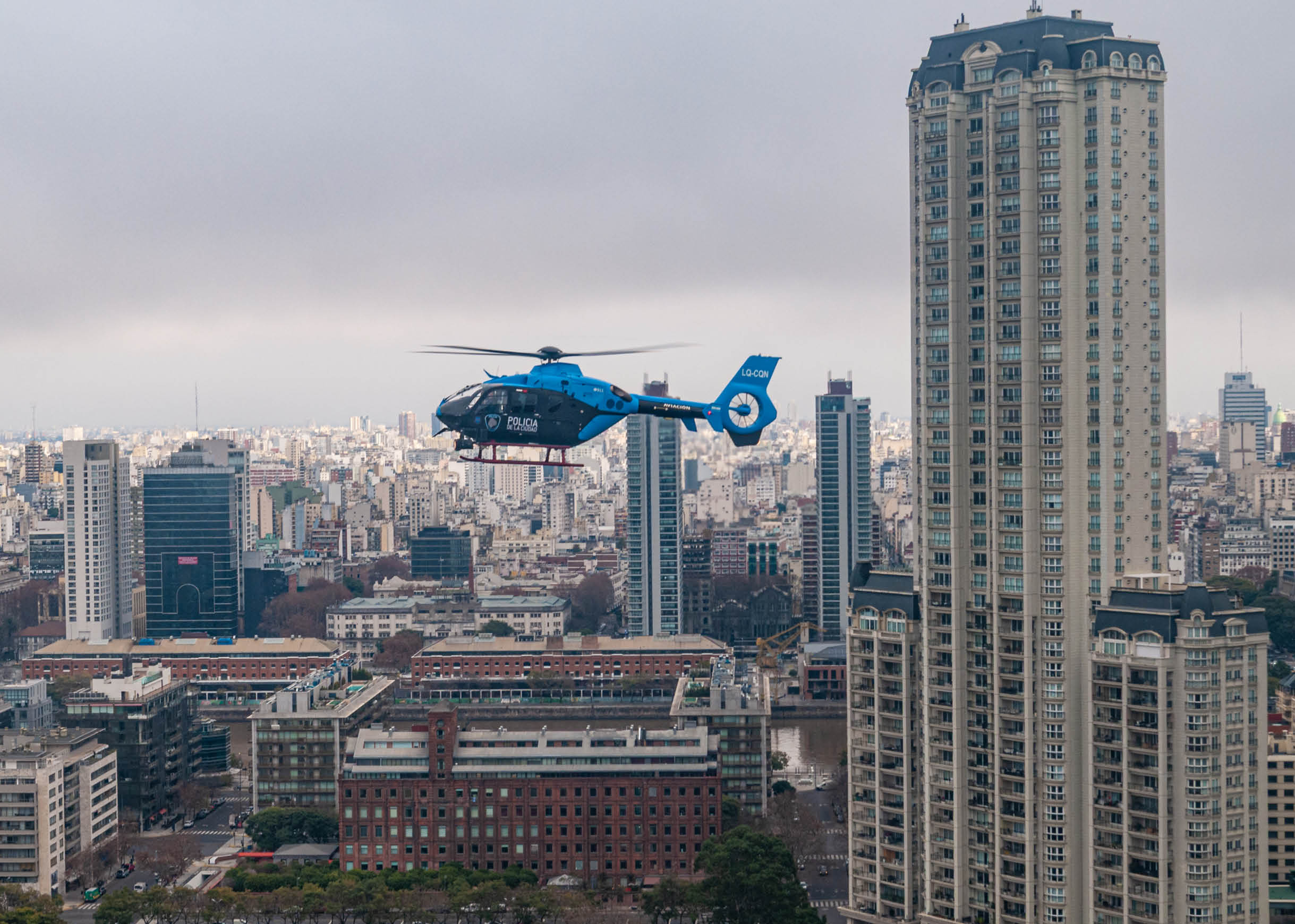 A blue helicopter is flying over a cityscape with tall buildings under a cloudy sky. Description generated by AI