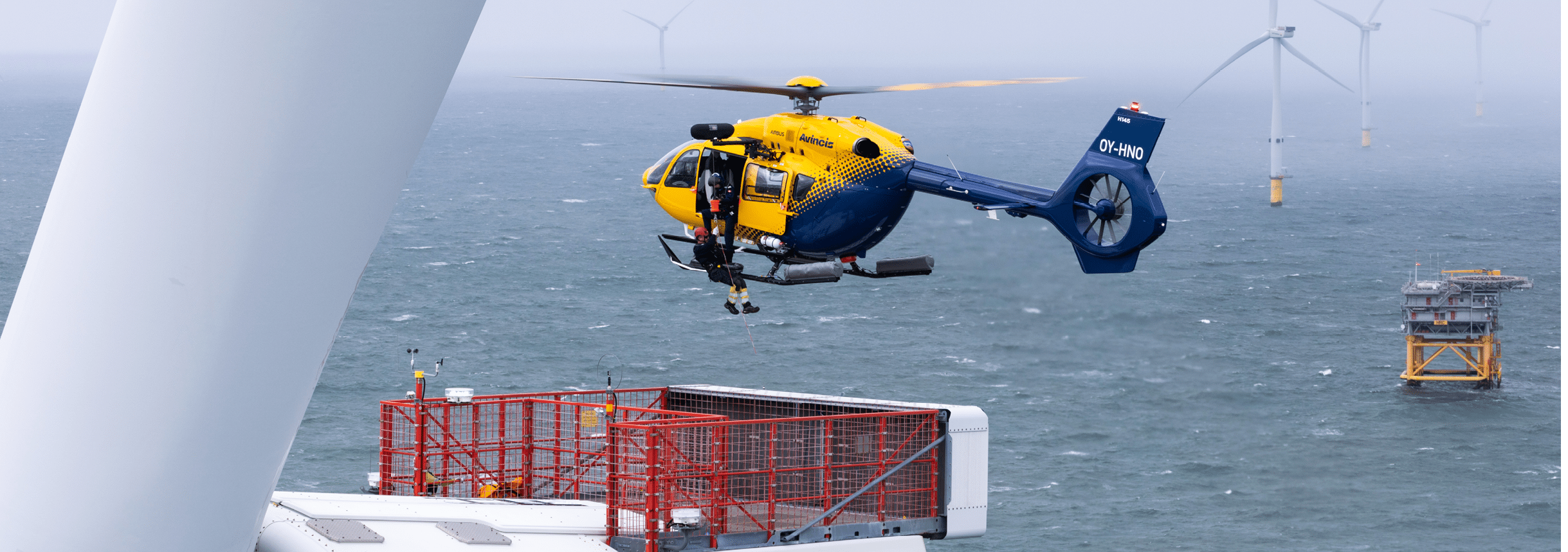 A helicopter hovers above an offshore platform with wind turbines visible in the background over a choppy sea. Description generated by AI