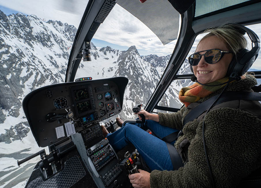 A person wearing sunglasses, headphones, and a warm jacket is smiling while piloting a helicopter over snowy mountainous terrain. Description generated by AI