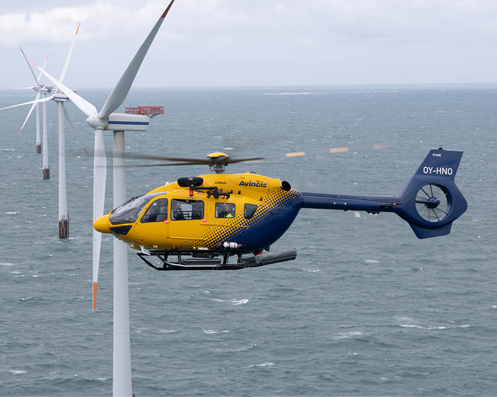 A yellow and blue helicopter flies near large offshore wind turbines above a choppy sea under a cloudy sky. Description generated by AI