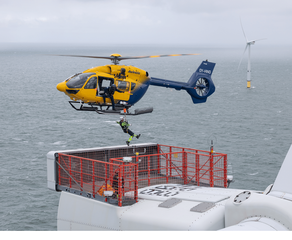 A person in safety gear is being lowered from a yellow and blue helicopter onto a platform on top of a wind turbine in a cloudy, overcast setting. Description generated by AI
