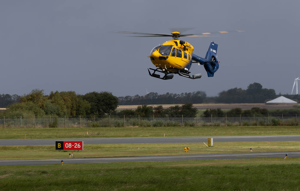 A yellow and blue helicopter is hovering above a grassy area near a runway with a cloudy sky and distant trees in the background. Description generated by AI