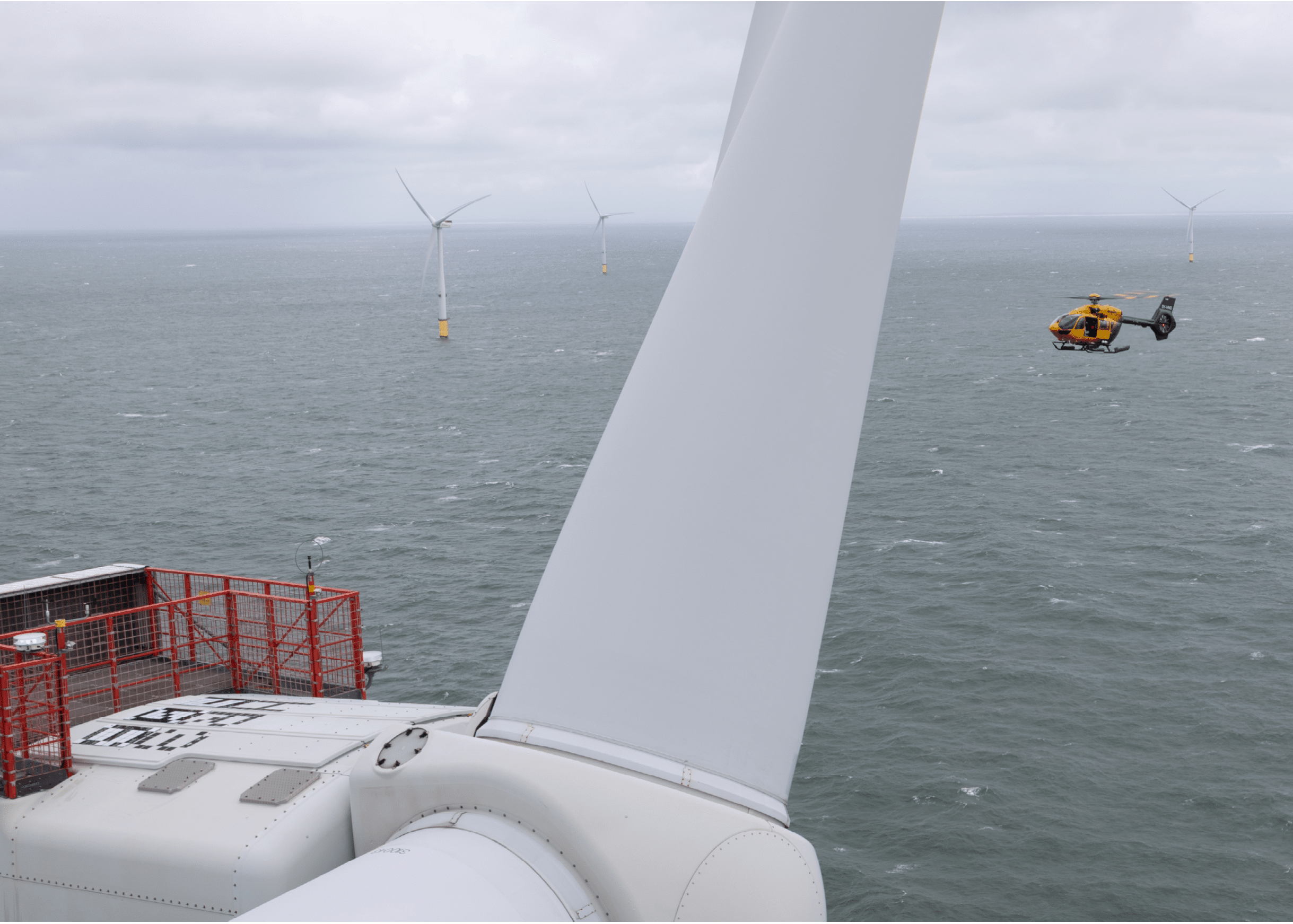 A helicopter flies near offshore wind turbines above a choppy sea under a cloudy sky. Description generated by AI