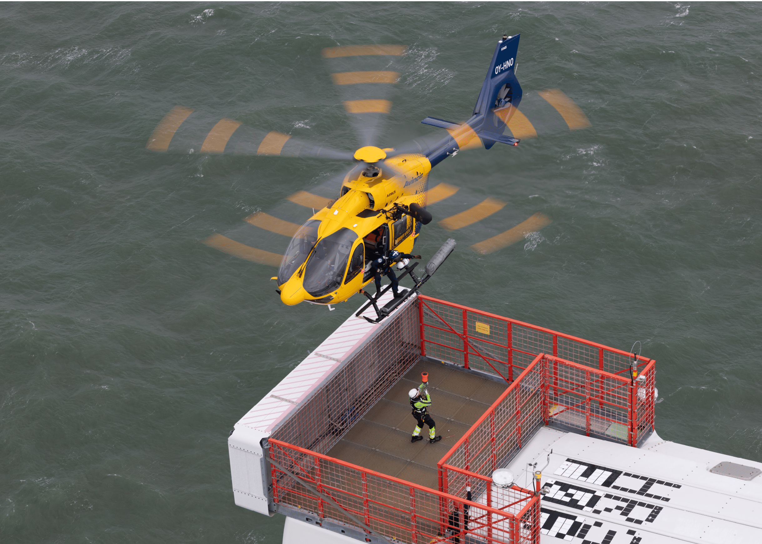A helicopter is landing on a platform above water where a person in safety gear is standing near a red railing. Description generated by AI