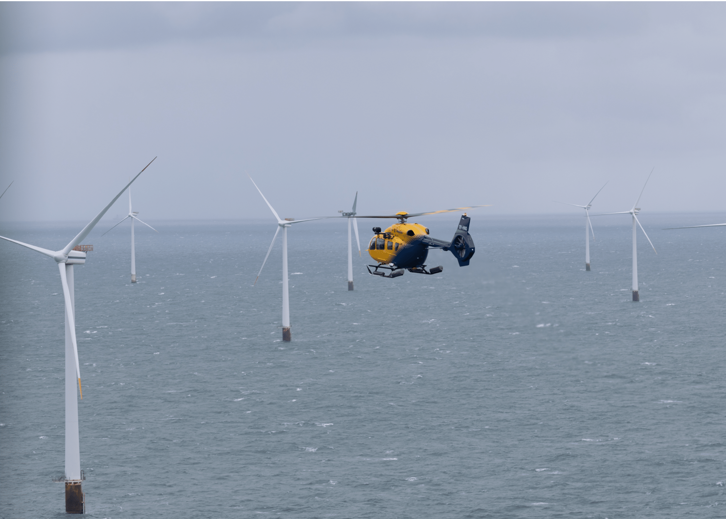A yellow and blue helicopter is flying over the ocean near several offshore wind turbines. Description generated by AI