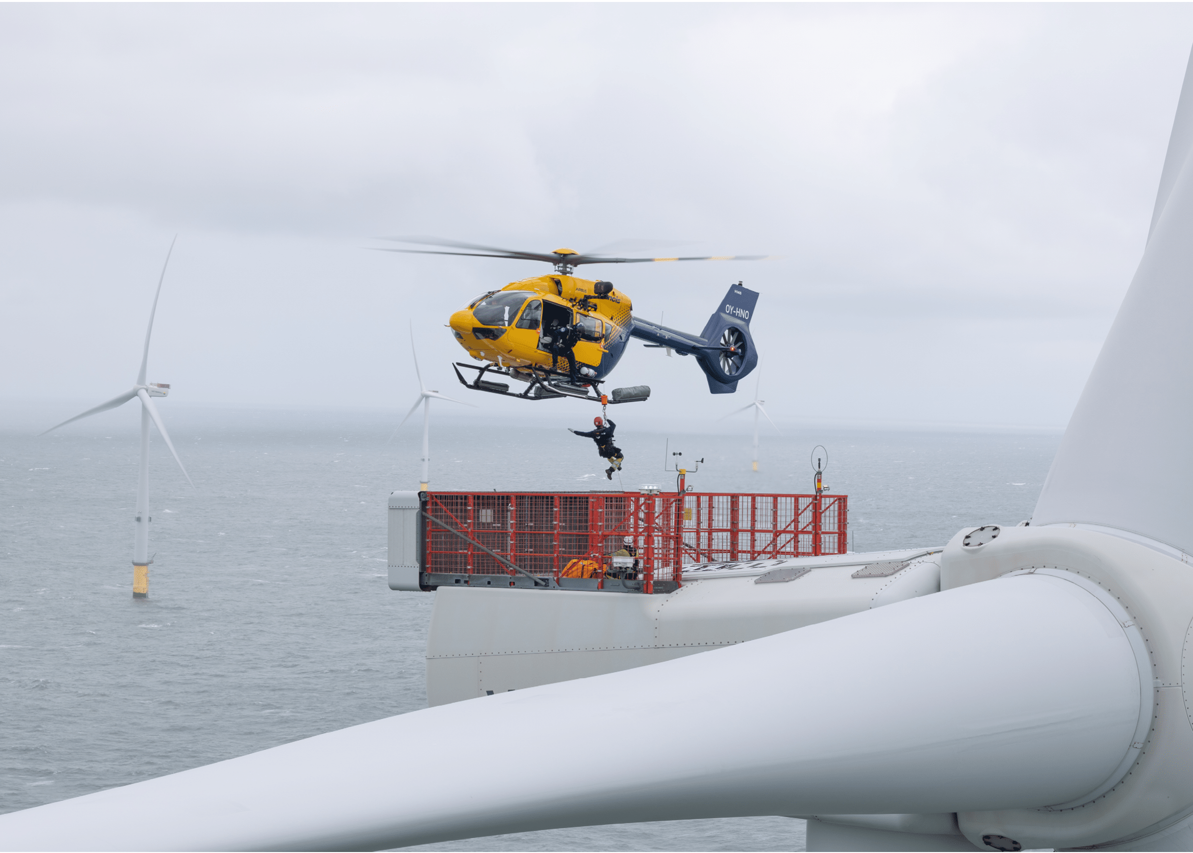 A yellow and blue helicopter hovers above an offshore wind turbine platform where a person is being lowered or lifted by a rope. Description generated by AI