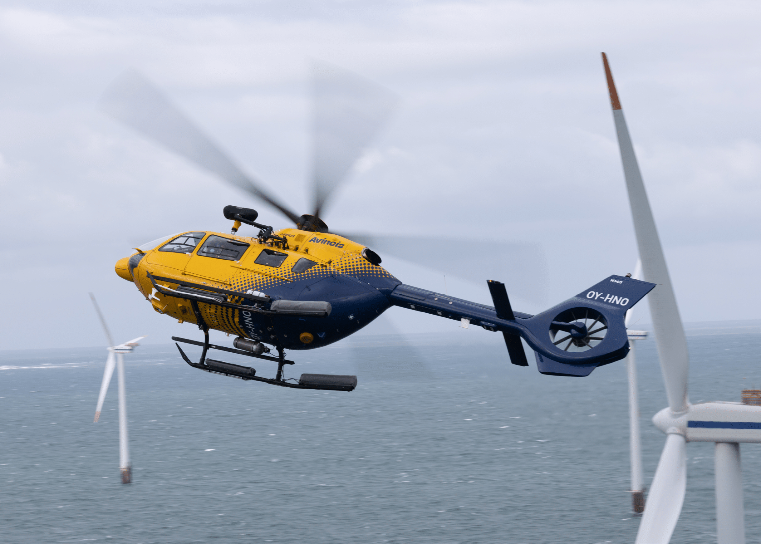 A close-up of a helicopter tail rotor with a wind turbine blade visible in the background against a cloudy sky. Description generated by AI