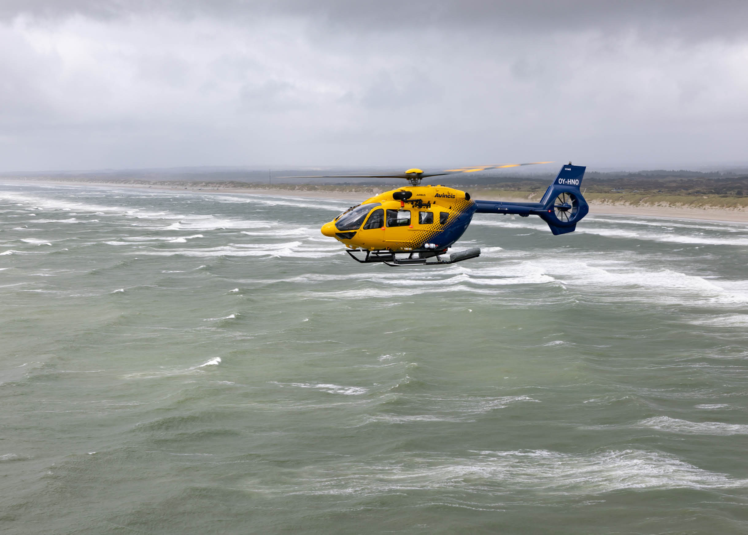 A yellow and blue helicopter is flying low over choppy ocean water near a sandy coastline under a cloudy sky. Description generated by AI